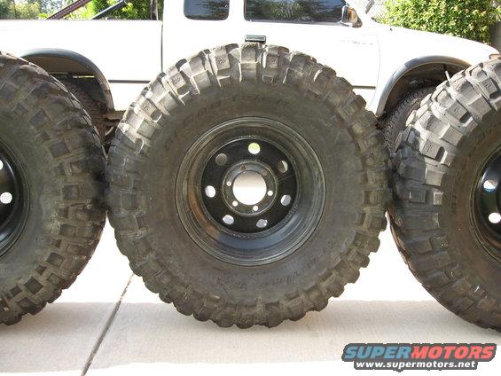 Three large off-road tires with aggressive tread patterns are lined up, with a white pickup truck visible behind them