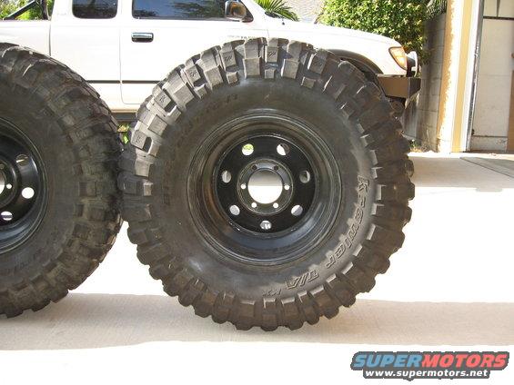 Two large off-road tires, one in focus, mounted on black rims, with a white pickup truck in the background