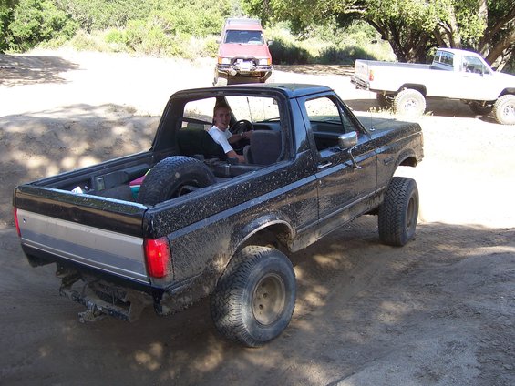 Black 1980s Ford Bronco with lifted suspension and spare tire in bed, driving on dirt road. Red SUV and white pickup truck in background