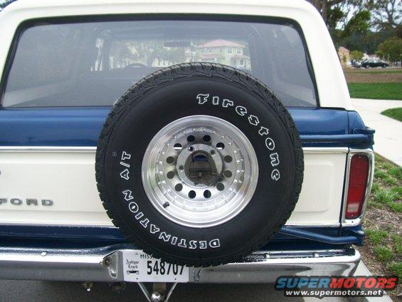 White and blue Ford Bronco rear with a spare tire mounted on the tailgate. Chrome bumper visible