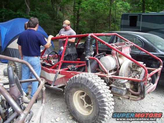 Red dune buggy with roll cage and large tires, parked next to a black car and a blue tent