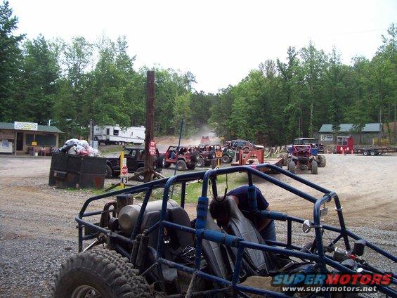 Several off-road buggies and ATVs parked in a dirt lot with trees in the background. A person is working on a blue buggy in the foreground