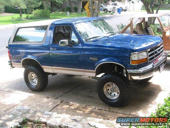 Blue 1996 Ford Bronco with lifted suspension and off-road tires parked on a driveway. Another vehicle is visible behind it