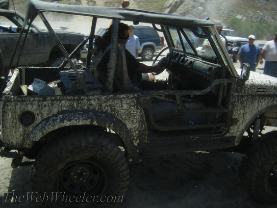 Muddy off-road vehicle with a roll cage, driver inside. Other vehicles and people in background
