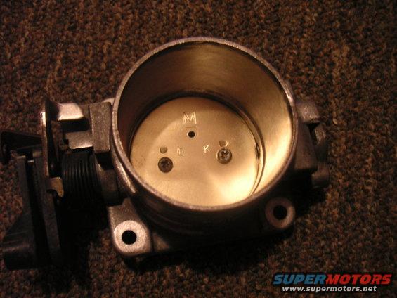 Throttle body with markings M, L, K, and D. It is mounted on a dark, textured surface