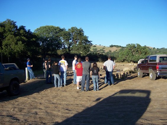People gathered around two SUVs in a dusty, rural area with rolling hills in the background