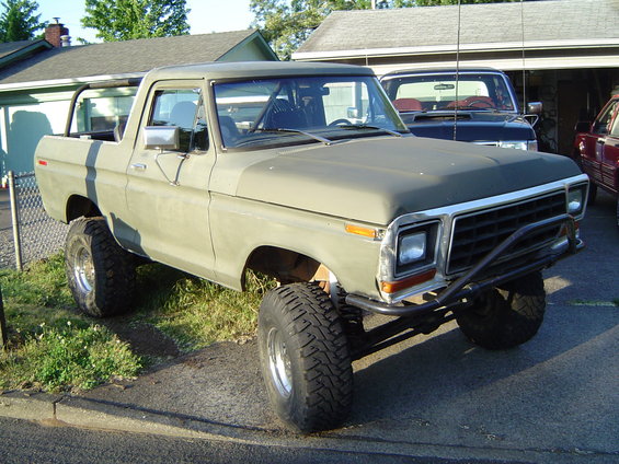 Olive green 1978 Ford Bronco with off-road tires and a brush guard parked in a driveway. Another truck is visible in the background