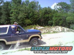 Blue and tan 1986 Ford Bronco driving on a dirt road with trees and sand dunes in the background