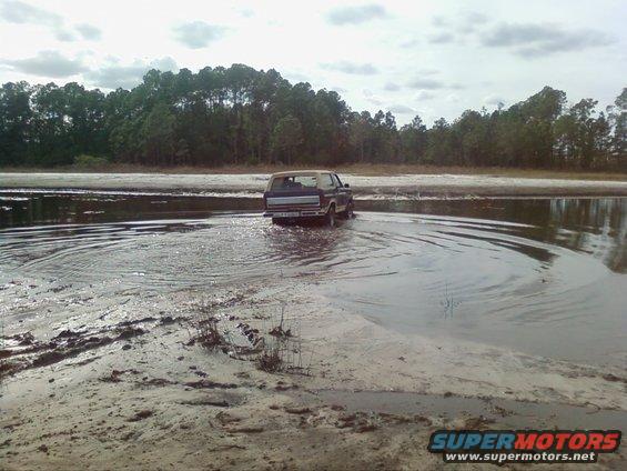 Ford Bronco driving through shallow water with trees in background