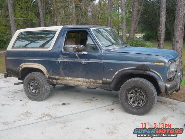 1986 Ford Bronco, blue, covered in mud, parked on concrete with trees in background