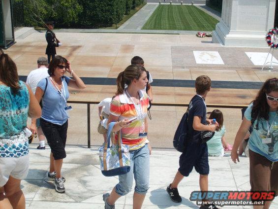 People walk up steps near a tomb guarded by a soldier. A grassy area is visible in the background