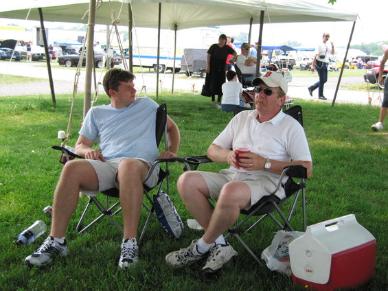 Two men relax in lawn chairs under a tent at an outdoor event. Cars are visible in the background