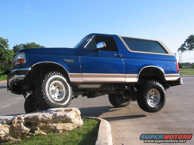 Blue Ford Bronco with tan trim, lifted on large tires, driving over a rock