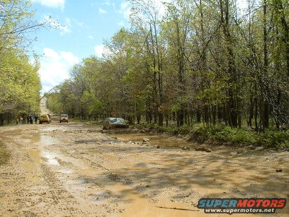 several vehicles driving on a very muddy and rutted dirt road through trees