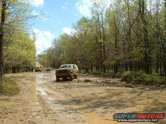 Muddy brown 1987 Ford Bronco driving through a flooded dirt road with trees on either side