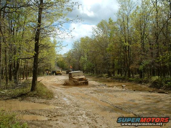 Several off-road vehicles navigate a muddy track through a wooded area