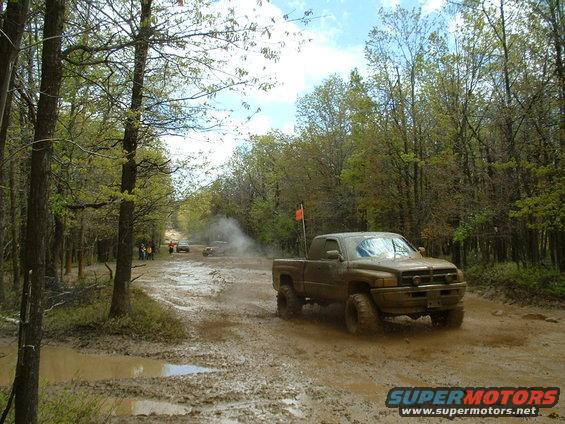 Muddy pickup truck driving through a wooded off-road course with other vehicles in the background