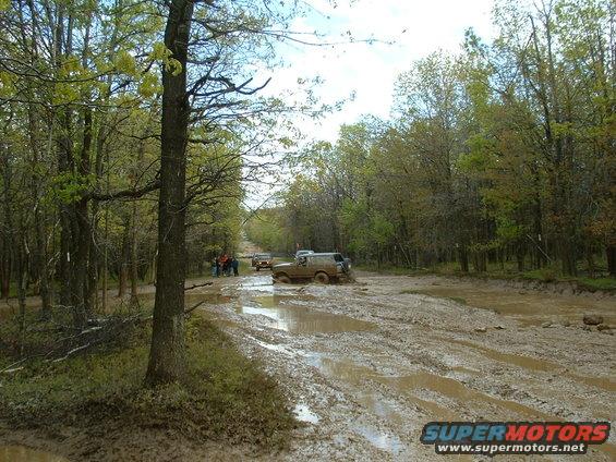 Several SUVs driving through a muddy, wooded trail
