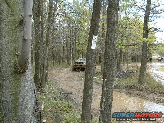 Muddy off-road trail with several SUVs driving through puddles and mud, surrounded by trees