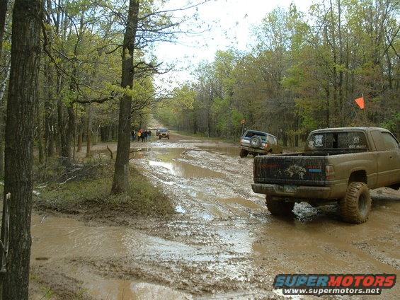 Muddy pickup truck rear view on a dirt road with trees and other vehicles