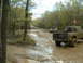1991 Ford Bronco - Several off-road vehicles navigate a muddy track through a wooded area.