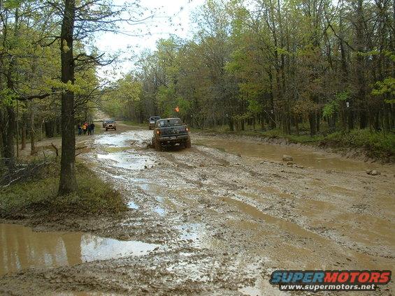 Several off-road trucks navigate a muddy trail with trees on either side