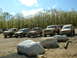 1991 Ford Bronco - A lineup of tan off-road vehicles, including a 1991 Ford Bronco, are parked on a dirt lot with trees in the background.