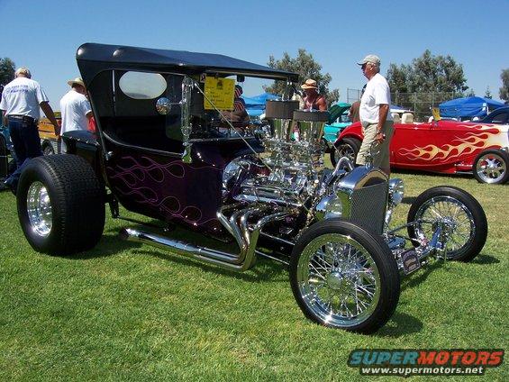 Purple hot rod with flame decals and a large V8 engine, parked on grass with other cars in background