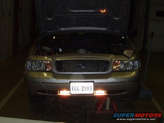 Gold Ford Crown Victoria with hood open, front view. Aftermarket lights installed below license plate