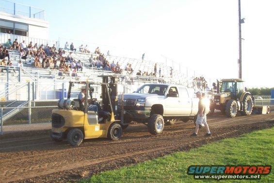 Forklift lifts front of pickup truck while tractor pulls it backward. Spectators watch from bleachers