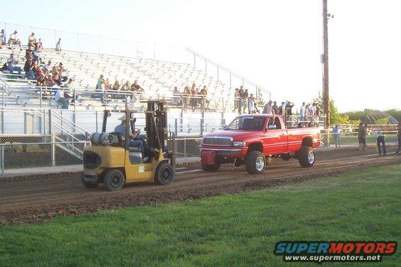 Red pickup truck with lifted suspension and large tires, being pulled by a yellow forklift on a dirt track in front of bleachers