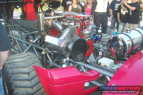 Close-up of a drag racing truck's engine and large tires. People watch in the background