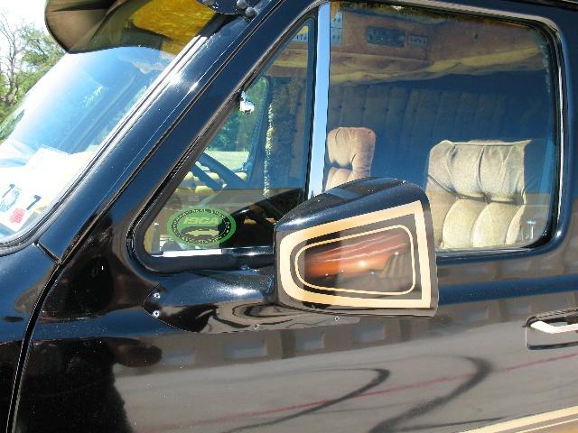 Black van side mirror with gold trim, showing reflection of amber turn signal. Interior has plush tan seats and wood-paneled walls