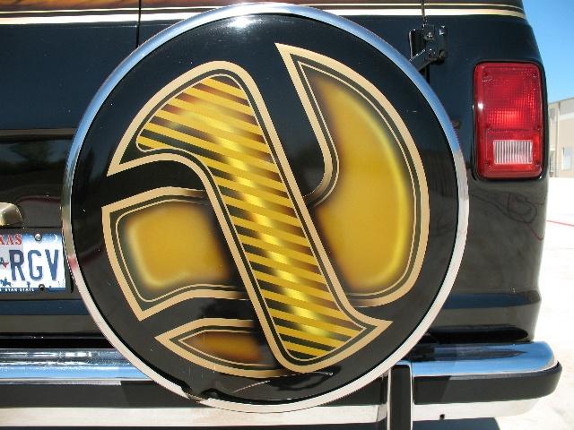 Rear view of a black van with a custom spare tire cover a yellow and black graphic. Texas license plate visible