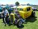 2002 Ford Mustang - A man sits on the fender of a bright yellow hot rod with flame decals and a prominent supercharger at an outdoor car show.