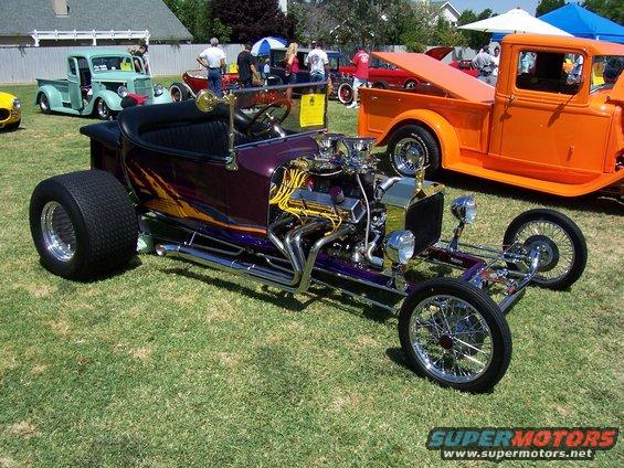 Purple custom hot rod with large rear tires and exposed V8 engine, parked on grass next to an orange pickup truck