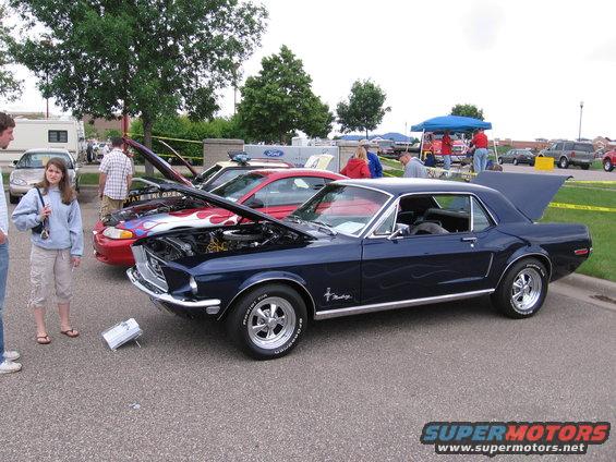 Dark blue classic car with flame decals, hood open, parked at a car show. Other cars and people are visible