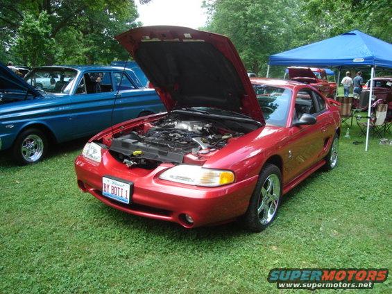 Red Ford Mustang with hood open, parked on grass next to a blue car. Another tent and cars are in the background