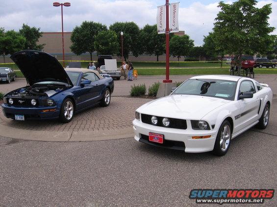 White Ford Mustang parked next to a blue convertible Mustang with its hood up
