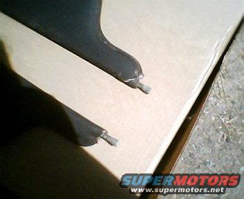Close-up of two black car parts with metal pins, resting on a light brown surface. Supermotors.net logo visible