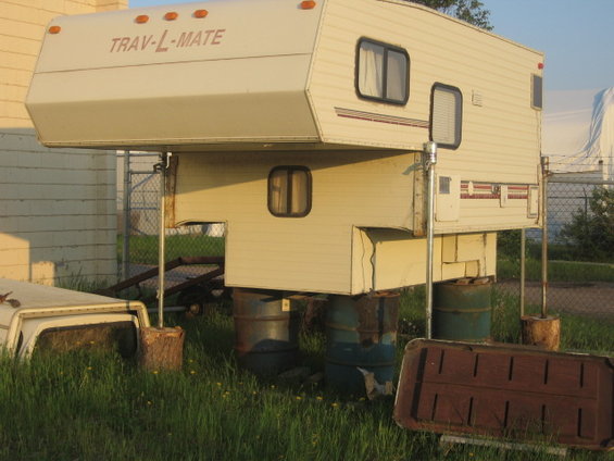Slide-in truck camper labeled TRAV-L-MATE, supported by barrels and wood