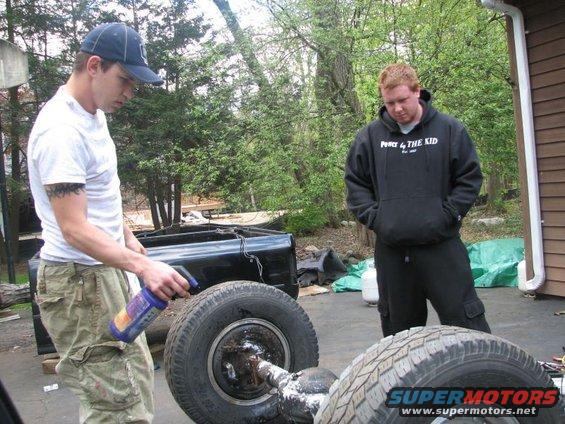 Two men are working on a truck bed and axle assembly outdoors. One man sprays a fluid onto the axle
