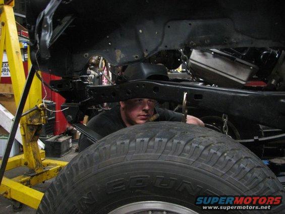 Mechanic peers under vehicle frame with large tire in foreground