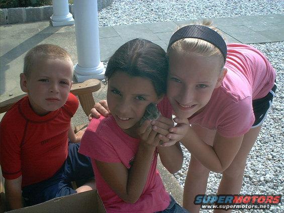 Three children, two girls and a boy, holding a small rodent-like animal outdoors