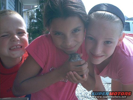 Three kids crowded together, one holding a small rodent. A white vehicle is visible in the background