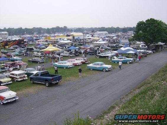 Outdoor car show with rows of classic cars and tents under a cloudy sky