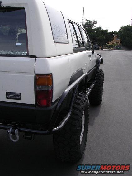 Rear quarter view of a white Toyota 4Runner with large off-road tires and a roll cage. It has a black bumper and fender flares