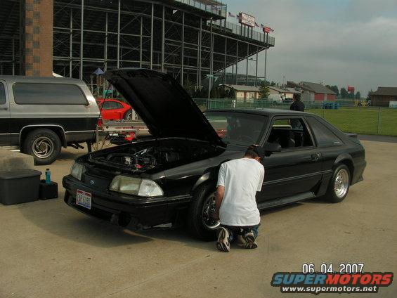Black coupe with hood open, person kneeling by front wheel. Silver SUV parked to the left. Grandstands in background