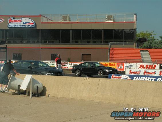 Black 2000 Ford Mustang GT parked on drag strip next to another car, Summit Motorsports Park in background