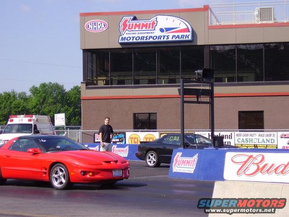 Red sports car and black sports car on drag strip in front of Summit Motorsports Park building. Ambulance in background
